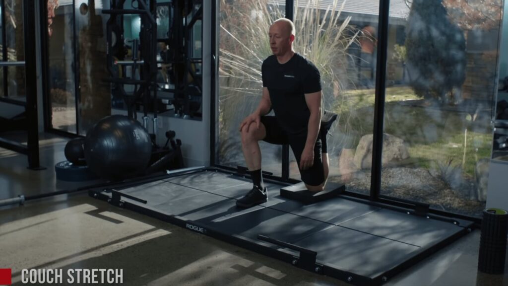 Chad shows the couch stretch. His right leg is bent at 90-degrees with his foot on the ground. The other leg is bent against a glass wall.